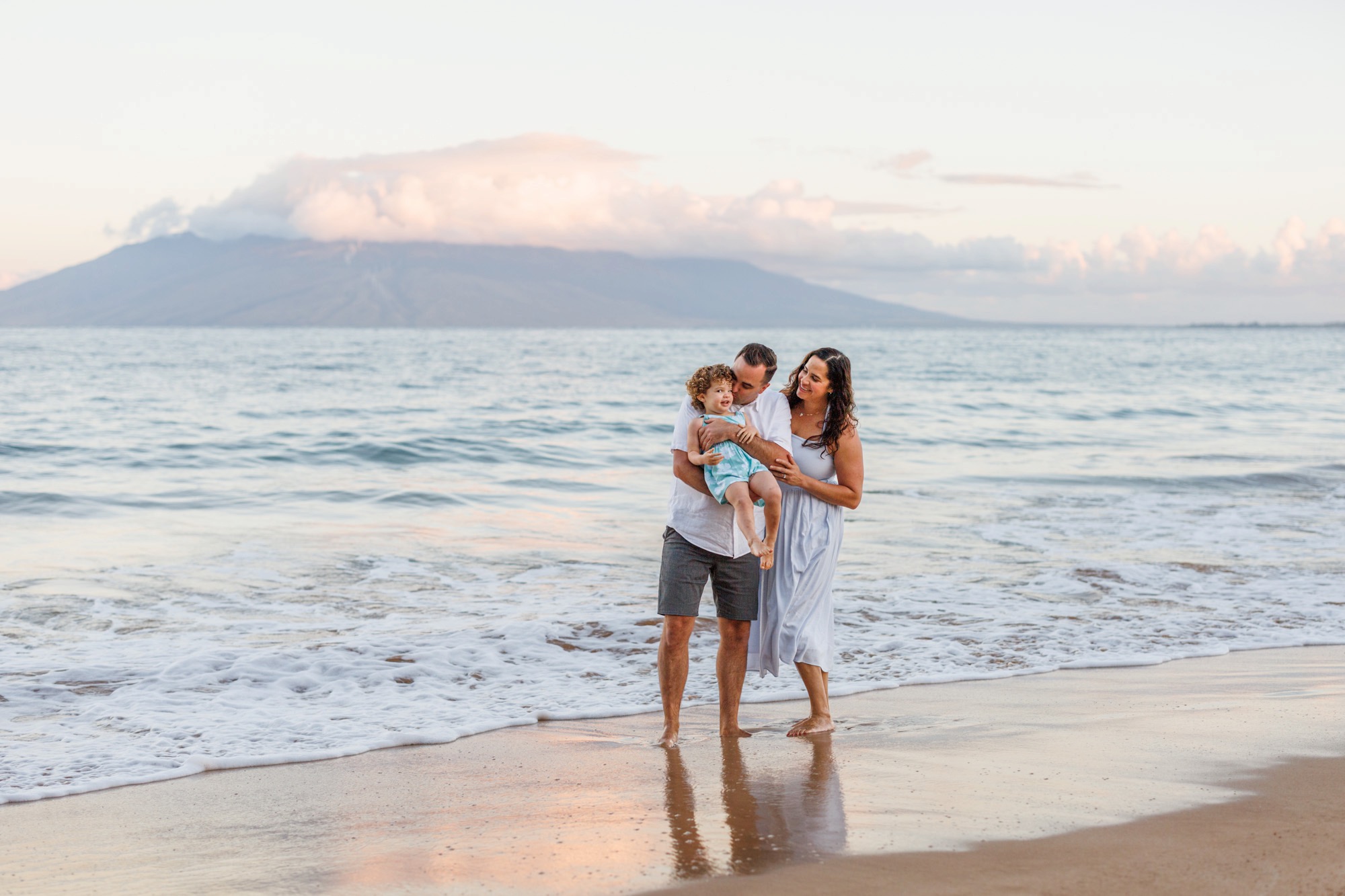 A family in white and soft blue embracing in the surf at sunrise in Wailea, with the West Maui Mountains behind